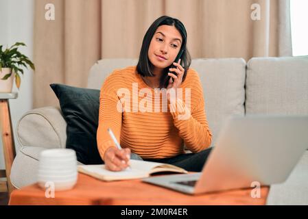 Its a good day to get things done. a young woman using a smartphone and writing notes while working on the sofa at home. Stock Photo