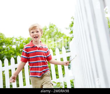 Energetic and excited in his backyard. A cute young boy running a stick along a white picket fence in his backyard. Stock Photo