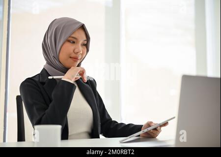 Attractive and professional Asian Muslim businessman or female manager in hijab using laptop, looking at laptop screen, working in the office. Stock Photo