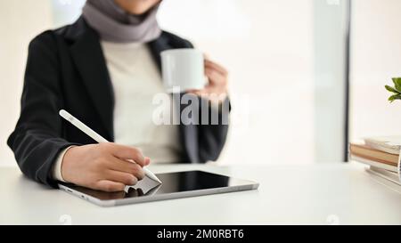 close-up shot of a professional Muslim businesswoman or female boss using digital tablet while sipping coffee at her desk. Stock Photo
