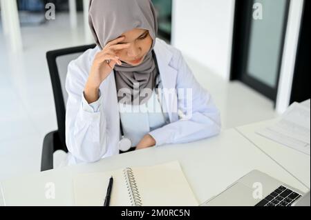 Stressed Asian Muslim female doctor concentrating on her medical research, working at her desk in the doctor's office. Stock Photo