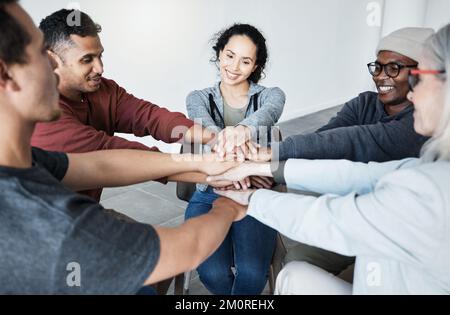 Diverse group of people sitting together in a circle and stacking their hands in the middle after therapy. Smiling support group celebrating after a Stock Photo