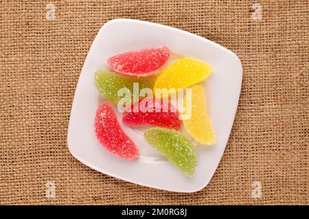Several sweet marmalade candies with white ceramic plate on jute cloth, macro, top view. Stock Photo