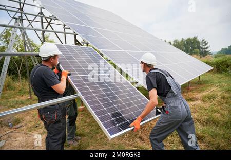 Workers installing solar panel on metal beams in field at daytime. Men wearing workwear and helmets. Ecological energy. Photo-voltaic collection of modules. Array - system of photo-voltaic panels. Stock Photo