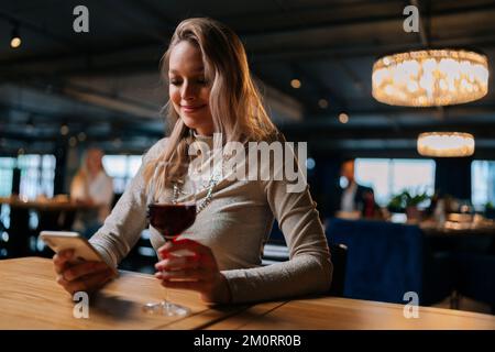 Low-angle view of smiling blonde young woman using smartphone, typing online message sitting at table holding in hand glass of red wine at restaurant. Stock Photo