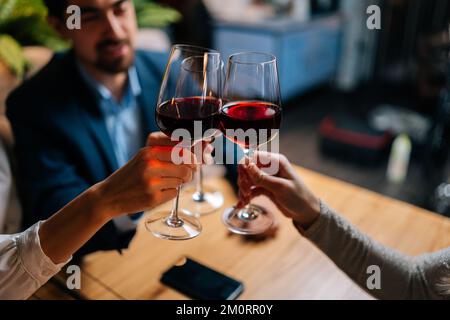 Close-up cropped shot of unrecognizable man in suit and two pretty women clinking glasses with red wine during festive dinner at table in restaurant Stock Photo