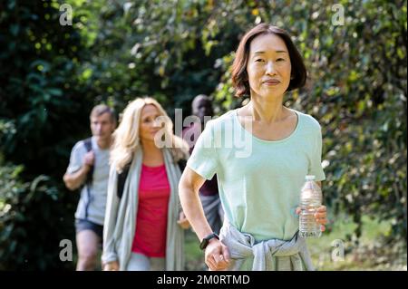 Senior Asian-American woman hiking in mountain trail with group of friends Stock Photo