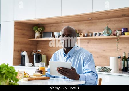 Senior African-American man sitting in kitchen eating fruit for breakfast while checking messages on digital tablet Stock Photo