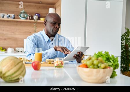 Senior African-American man sitting in kitchen eating fruit for breakfast while checking messages on digital tablet Stock Photo