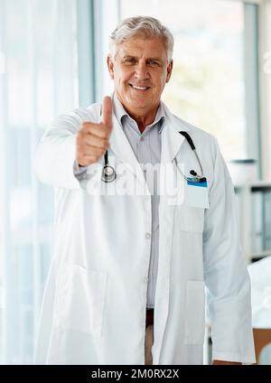 Happy mature caucasian male doctor smiling while showing a thumbs up working at a hospital alone. One senior man wearing a labcoat standing and Stock Photo