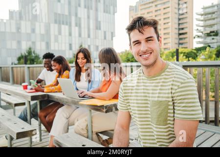 On foreground a positive and smiling young student guy looking at camera sitting outside at the University Campus. Meeting of friends group laughing and connecting to the internet using their laptops outside on high school. Education concept. High quality photo Stock Photo