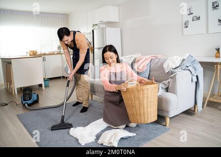 Young Asian couple doing houserkeeping together at home. Beautiful wife doing laundry and husband doing vacuum cleaning on the floor. Housework and chores concept Stock Photo