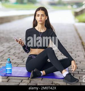 Beautiful young woman leading a healthy lifestyle, listening to music while sitting on a gymnastic mat, resting after playing sports on the street Stock Photo