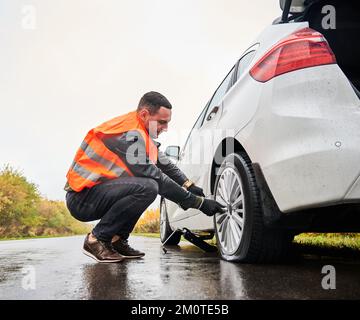 Male auto mechanic in work vest unscrewing lug nuts on car wheel in process of new tire replacement. Young man using wrench while changing flat tire on the road. Concept of emergency road service. Stock Photo