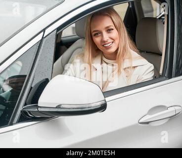 Portrait of pretty smiling blonde woman sitting behind the steering wheel in automobile. Happy lady resting in driver's seat inside her comfortable white car. Stock Photo