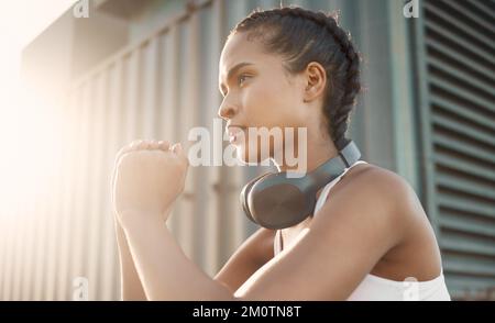 One fit young hispanic woman wearing headphones and doing squat exercises while training in an urban setting outdoors. Focused female athlete doing Stock Photo