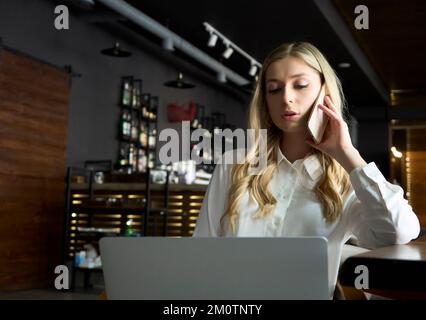 Joyful elegant female operator of call center is looking at the camera, smiling. Intelligent business woman is sitting at the table in cafe, working Stock Photo