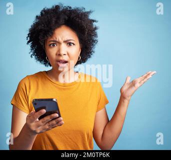 What does this text even me. a young woman standing against a blue background in the studio and looking confused while using her cellphone. Stock Photo