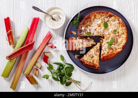 gluten free swiss rhubarb tart, rhabarberwahe, sprinkled with sliced almonds and icing sugar on white platter on white wooden table with ingredients, Stock Photo