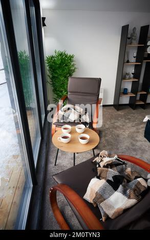 Breakfast with cups of tea on wooden table and cozy chairs in nice decorated room inside. Small coffee table with mugs and armchairs with checkered plaids next to window in winter. Concept of comfort. Stock Photo
