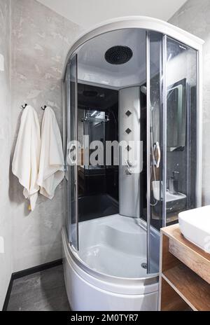 Small gray tile bathroom with fully equipped shower cabin and ceramic sink indoor. Front view of modern small shower stall with two white towels next in washroom. Concept of shower equipment. Stock Photo