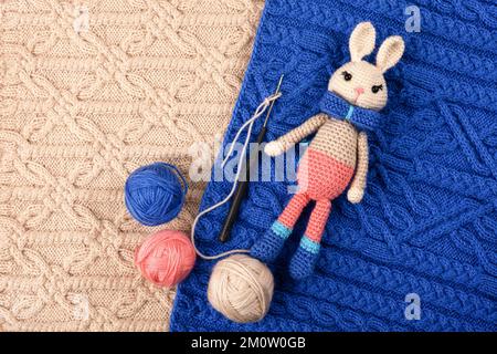 Knitted children's toy hare or bunny with a balls of thread and a crochet hook on a knitted blue and beige cloth. The symbol of 2023. Stock Photo