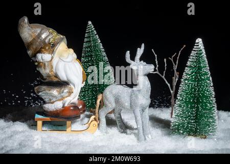 Creative Christmas card. A toy shining silver deer carrying a sleigh with Santa Claus with gifts through the snowy forest among the Christmas trees at Stock Photo