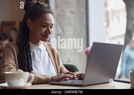 Adorable African woman student types text on laptop keyboard, works, studies, doing research or prepares for exam online Stock Photo