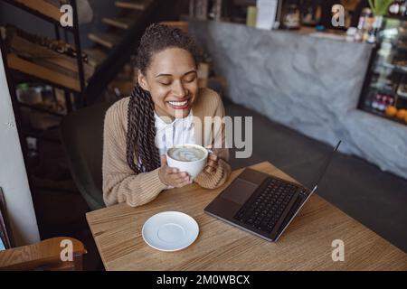 Delightful young African American woman, student, blogger, freelancer holding cup with cappuccino drink and enjoying coffee break while studying remot Stock Photo