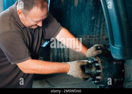 Auto mechanic repairs hub of truck in car repair shop. White Caucasian man in gloves disassembles car. Service for repair of large trucks and tractors. Background. Real workflow.. Stock Photo