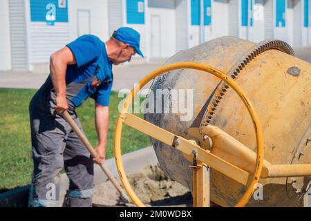 Uniformed construction worker works on construction site on summer day. Elderly bricklayer shovels cement and sand into concrete mixer. Authentic workflow. Background.. Stock Photo