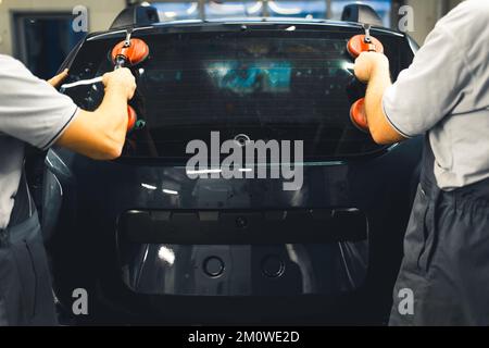 Back indoor view of two mechanics in gray uniforms using suction cups to remove rear window from a black car. Professional workers. High quality photo Stock Photo