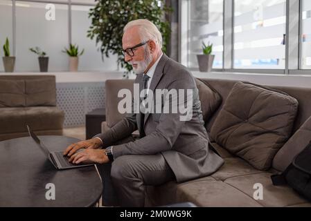 Senior businessman using laptop while sitting on sofa at train station lobby. Stock Photo