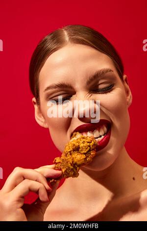 Young woman with red lipstick eating fried chicken, nuggets over vivid red background. Fast food lover. Food pop art photography. Stock Photo