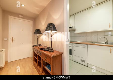 Entrance hall and housing distributor with a white wooden access door, a wooden sideboard with twin lamps and access to an elongated kitchen Stock Photo