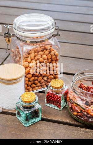 Transparent glass jars filled with peanuts, red pepper, dried chilli peppers, salt flakes on wooden surface Stock Photo