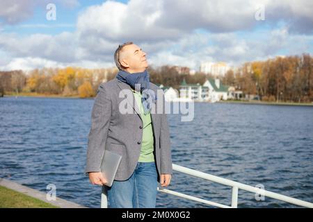 Portrait of happy middle-aged man standing with closed eyes on bank of river in city, enjoying sunlight, holding laptop. Stock Photo