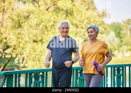 Older Asian and Caucasian wife and husband wear activewear strolling along bridge in summer park, enjoy morning sportive walk together outside. Health Stock Photo