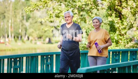 Asian and Caucasian senior wife and husband wear activewear strolling along bridge in summer park, enjoy morning sportive walk together outside. Healt Stock Photo