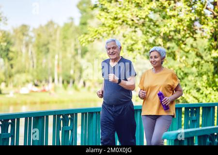 Asian and Caucasian middle-aged wife and husband wear activewear strolling along bridge in summer park, enjoy morning sportive walk together outside. Stock Photo