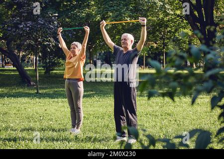 Senior 60 spouses wear sportswear doing hands exercises, training arms outdoor in summer park in the morning using resistance rubber bands. Healthy li Stock Photo