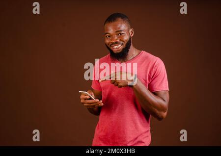 An attractive happy African man holding a phone and pointing at it isolated on a brown background. Stock Photo
