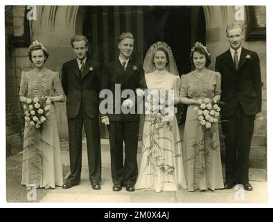 Original WW2 era wedding photograph group portrait of a happy pretty bride, wearing a white bridal gown, with puff sleeves and headdress with veil, bridegroom and best man in suits and attractive bridesmaids again wearing dresses with puff sleeves, standing outside a church, from London and Northern Studios, Alnwick, Northumberland, Northeastern England, U.K. Circa 1939. Stock Photo
