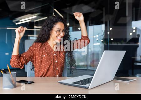 A beautiful young Latin American woman freelancer sits in the office at the table with a laptop, looks at the monitor, raised her hands, rejoices. Stock Photo