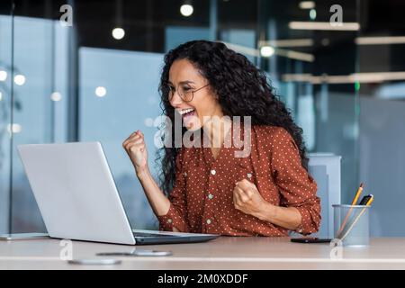 Happy beautiful young Latin American woman sitting in the office in front of the laptop monitor, happy about the win, good news, victory. He smiles, shows a yes gesture with his hands. Stock Photo