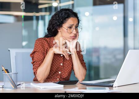 Burnout at work. Young Latin American business woman sitting tired and upset at the table in the office at the laptop, a lot of work, deadlines. Stock Photo