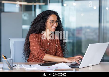 Online meeting. A young beautiful Latin American woman is sitting in the office at a desk, talking on a video call from a laptop. Working meeting, webinar, online conference Stock Photo
