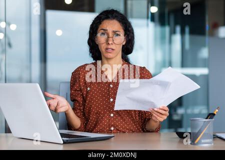 Worried young Latin American woman, student sitting in the library at a table with a laptop, studying. He holds papers, abstracts, documents in his hands. He looks tiredly at the camera. Stock Photo