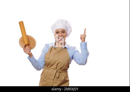 Delighted woman chef holding wooden board and rolling pin, points with finger at copy advertising space white background Stock Photo