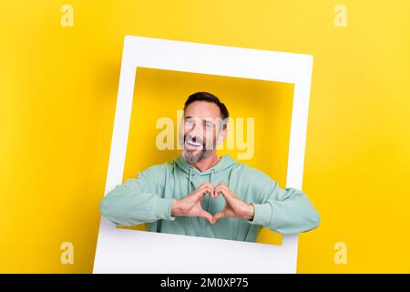 Portrait of satisfied positive man dressed green hoodie posing in white frame fingers show heart isolated on yellow color background Stock Photo